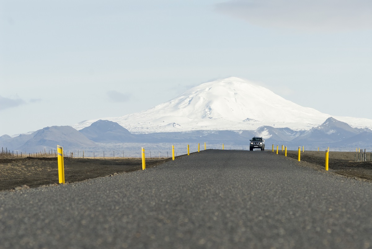 Een auto rijdt over de weg met op de achtergrond de besneeuwde Hekla vulkaan in IJsland.