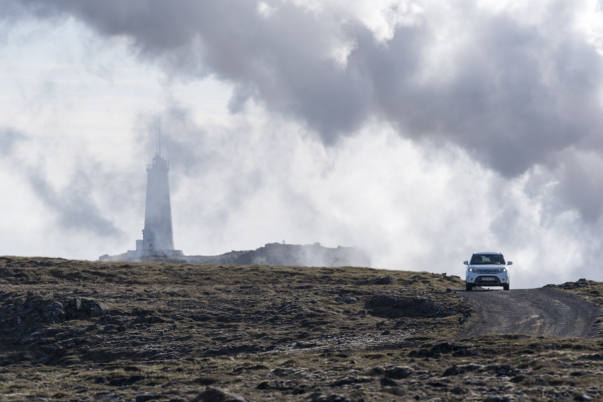 Een auto op een gravelweg in IJsland met op de achtergrond de Reykjanes vuurtoren in de wolken.