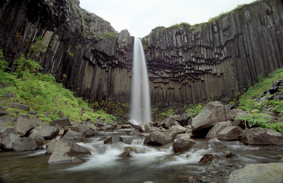 De waterval valt langs basalt rotsen naar beneden tussen groene varens