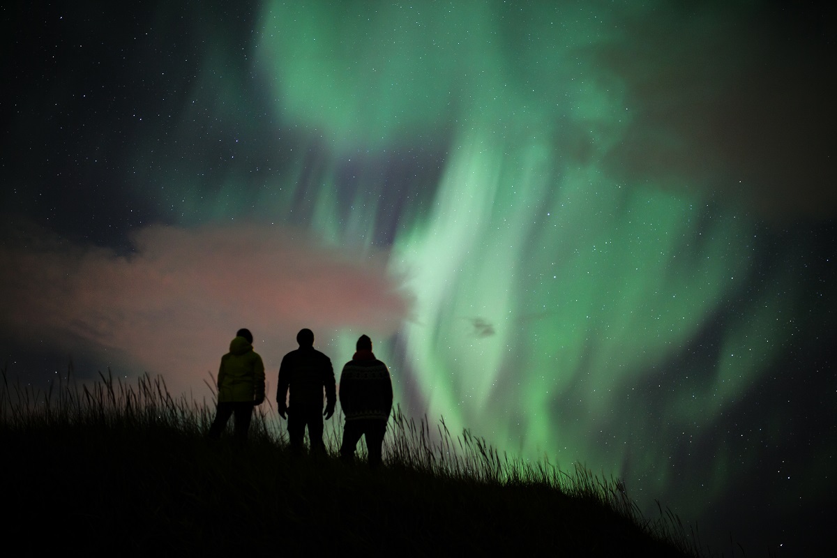 3 reizigers staan te kijken naar het groene noorderlicht dat boven hun hoofd aan het dansen is.